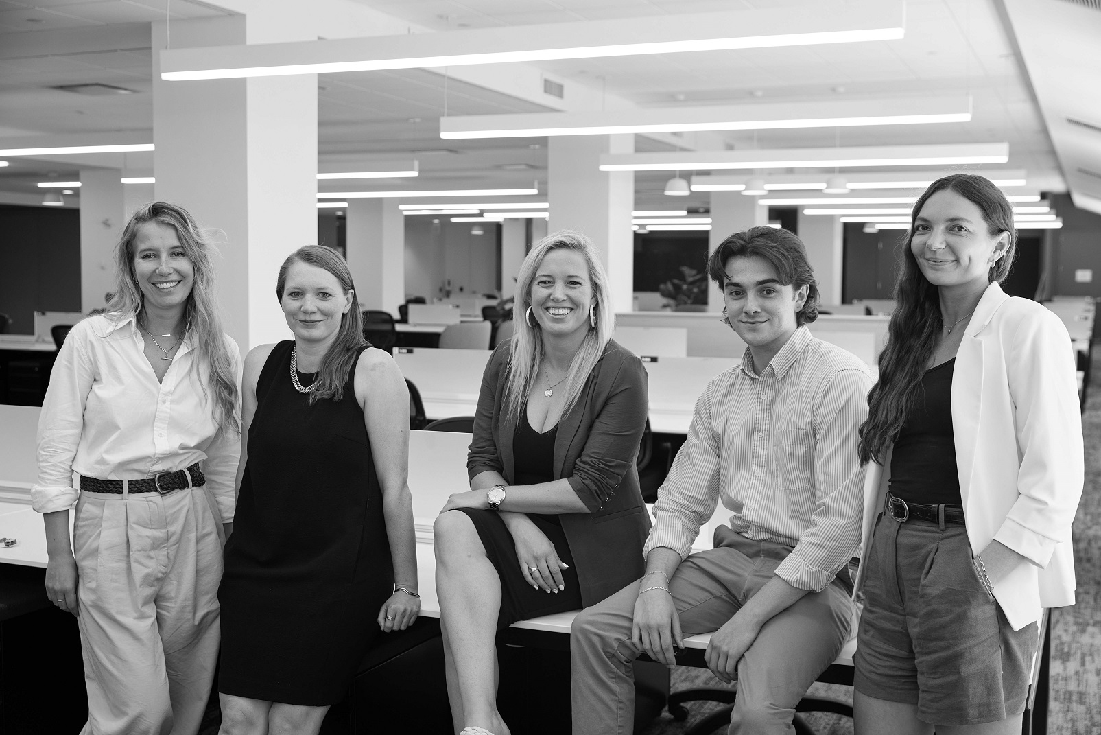Black and white photo of four women and one man leaning against a desk in an office